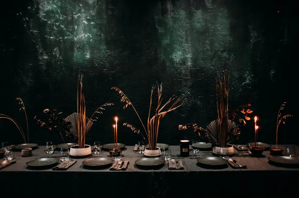 Mesa larga puesta con platos de piedra, velas encendidas y ramas secas contra una pared oscura