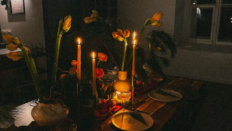 Velas encendidas y flores de estación en recipientes de bronce sobre una mesa oscura
