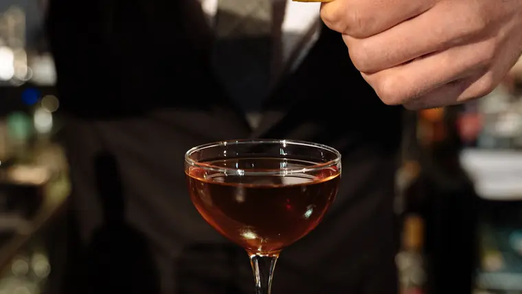 Bartender terminando un trago de autor en copa, con la barra en penumbra detrás