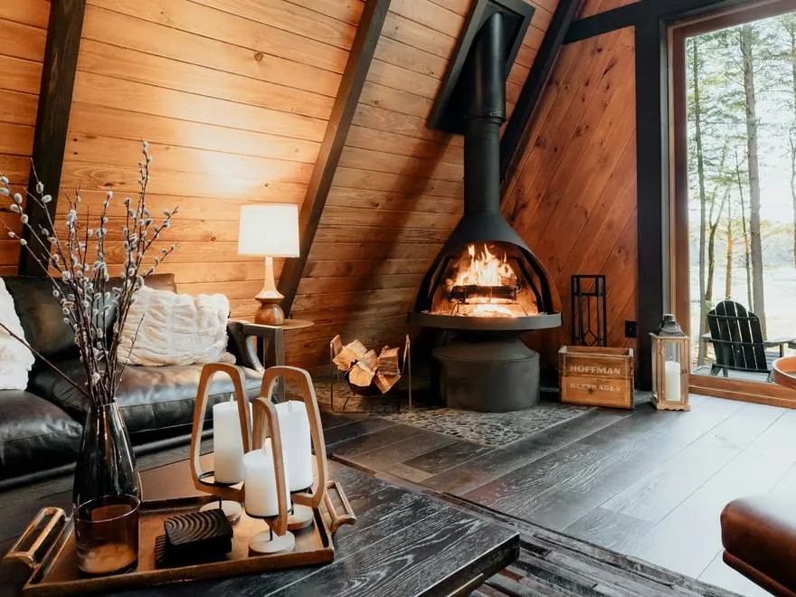 Living de una cabaña de madera con una estufa a leña moderna encendida y un ventanal con vista al bosque y al lago