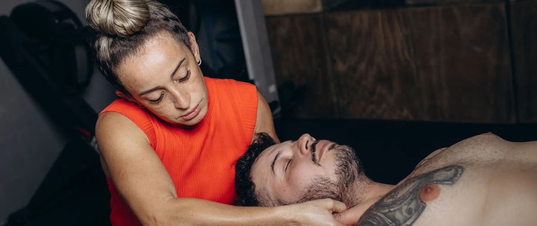 La kinesióloga, de remera roja, sosteniendo con las dos manos la nuca de un deportista recostado en la camilla, en un gimnasio de paredes grises